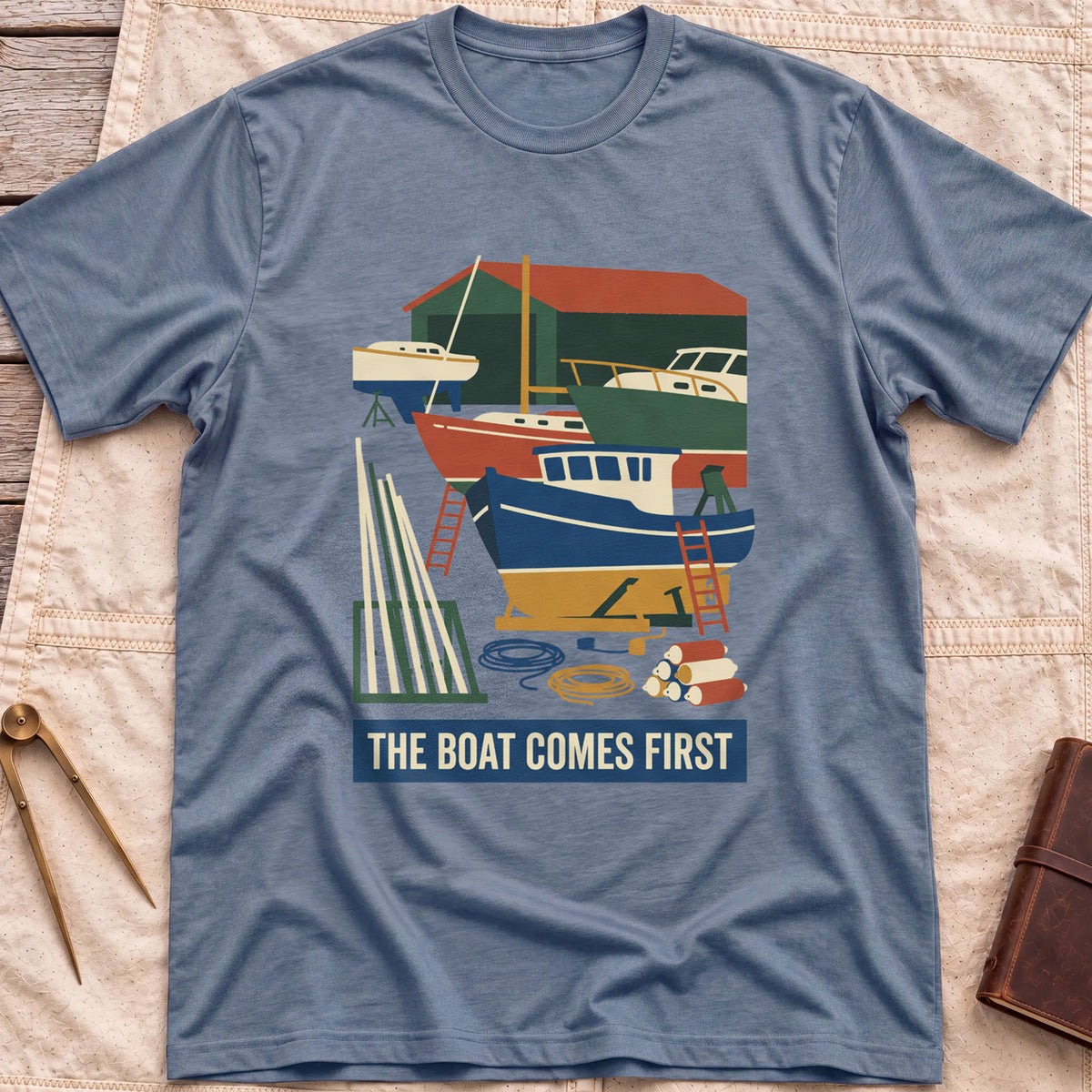 Front view of the heather indigo The Boat Comes First T-Shirt with a colourful boatyard scene of boats on stands, ladders, timber and coiled rope.
