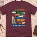 Front view of the maroon The Boat Comes First T-Shirt with a colourful boatyard scene of boats on stands, ladders, timber and coiled rope.