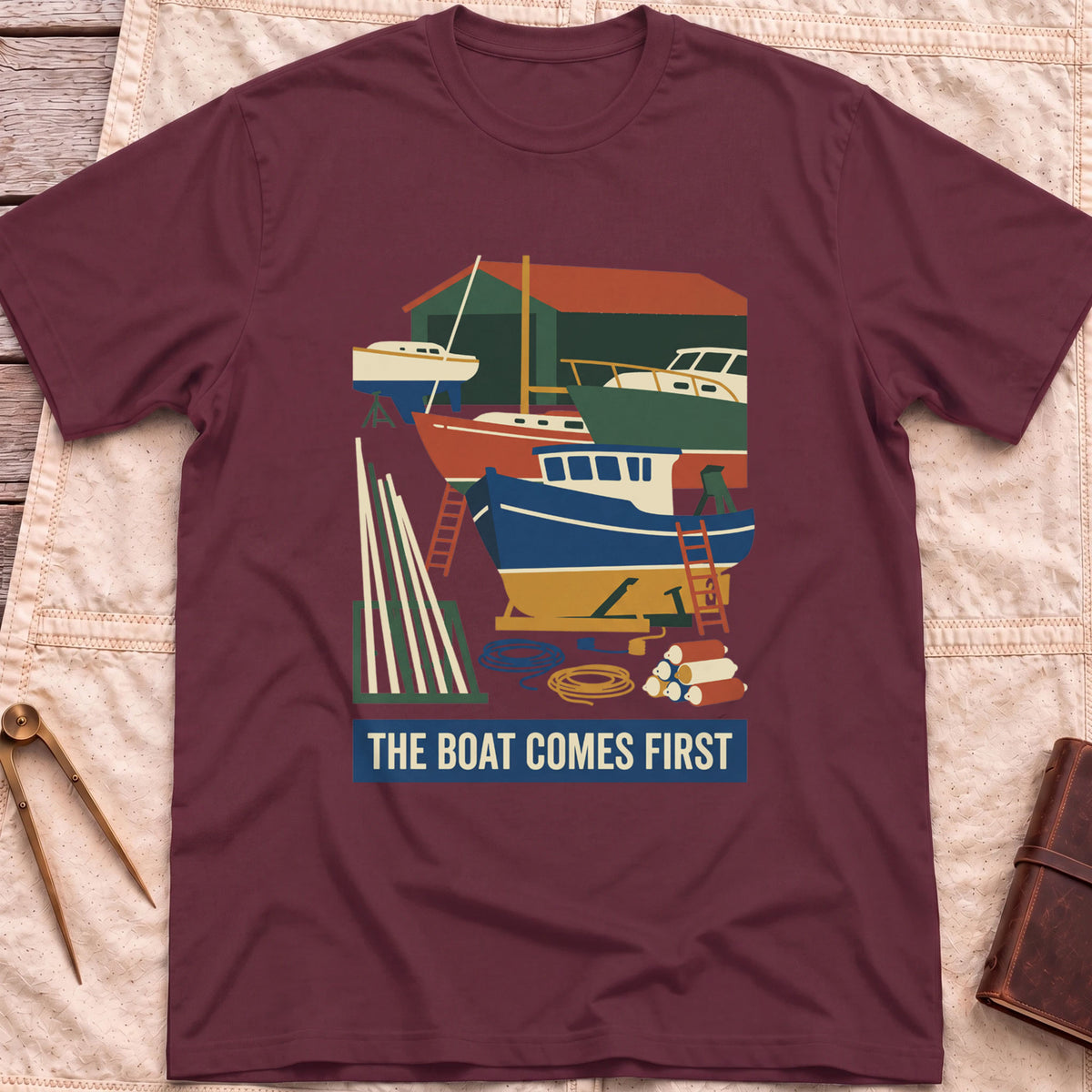 Front view of the maroon The Boat Comes First T-Shirt with a colourful boatyard scene of boats on stands, ladders, timber and coiled rope.