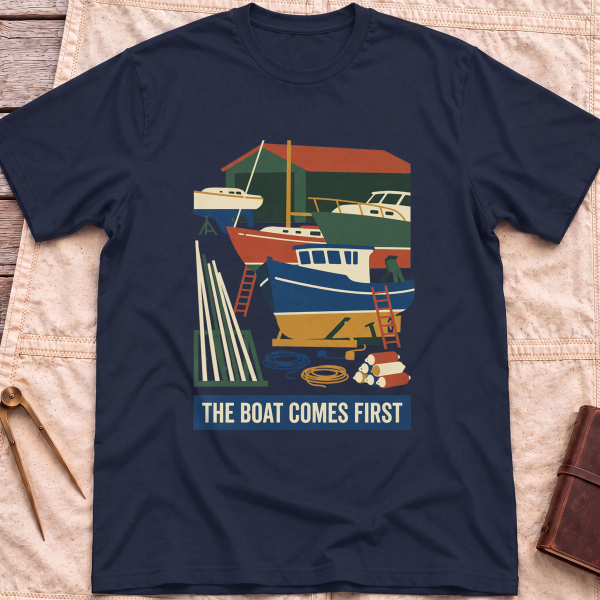 Front view of the navy The Boat Comes First T-Shirt with a colourful boatyard scene of boats on stands, ladders, timber and coiled rope.