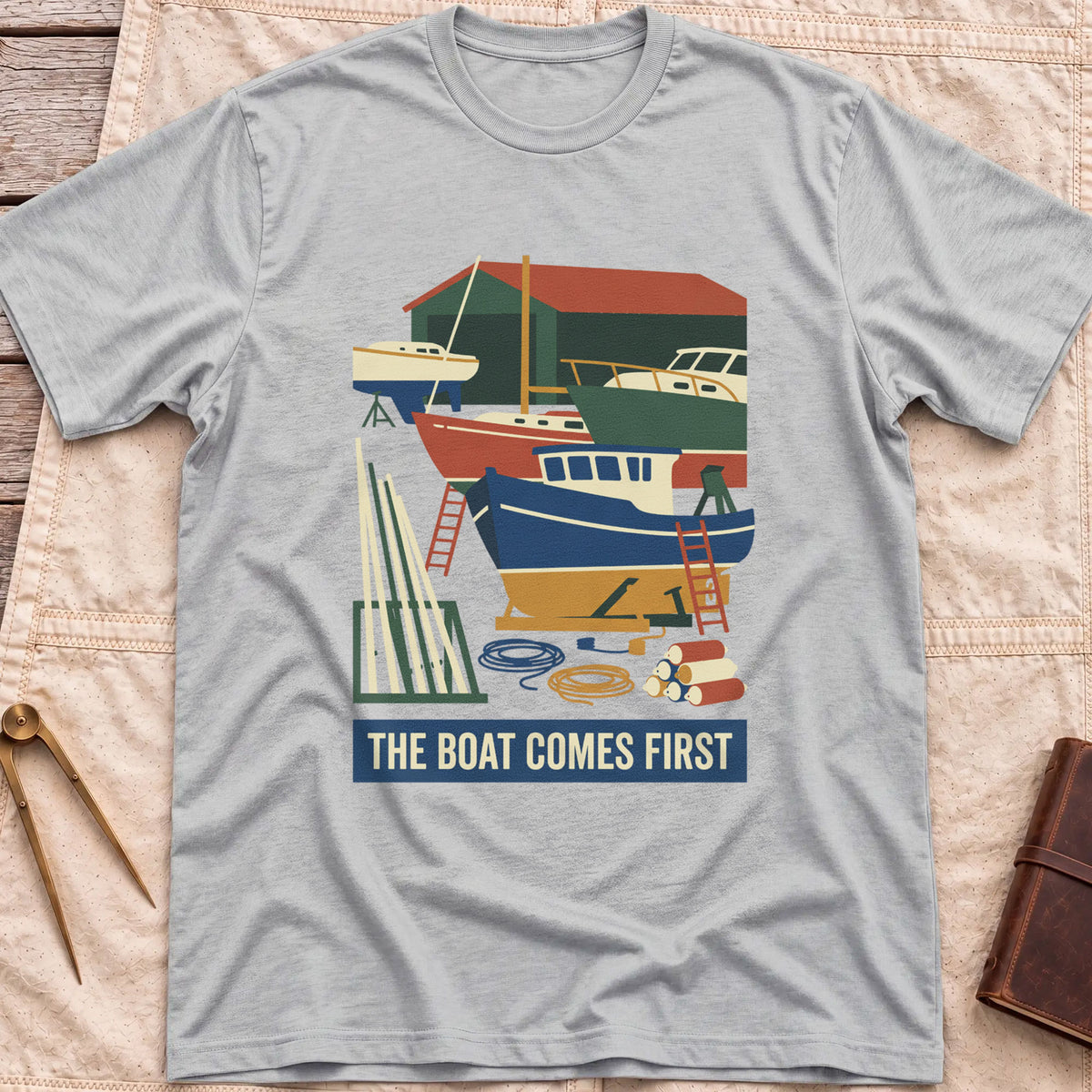 Front view of the sport grey The Boat Comes First T-Shirt with a colourful boatyard scene of boats on stands, ladders, timber and coiled rope.
