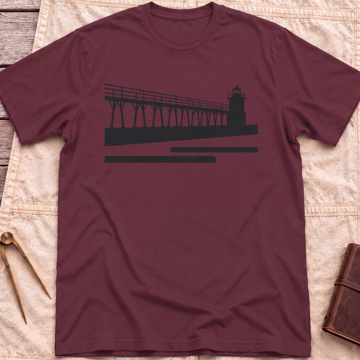 Front view of the maroon The Catwalk T-Shirt with a silhouette of a raised walkway running along a pier to a lighthouse.
