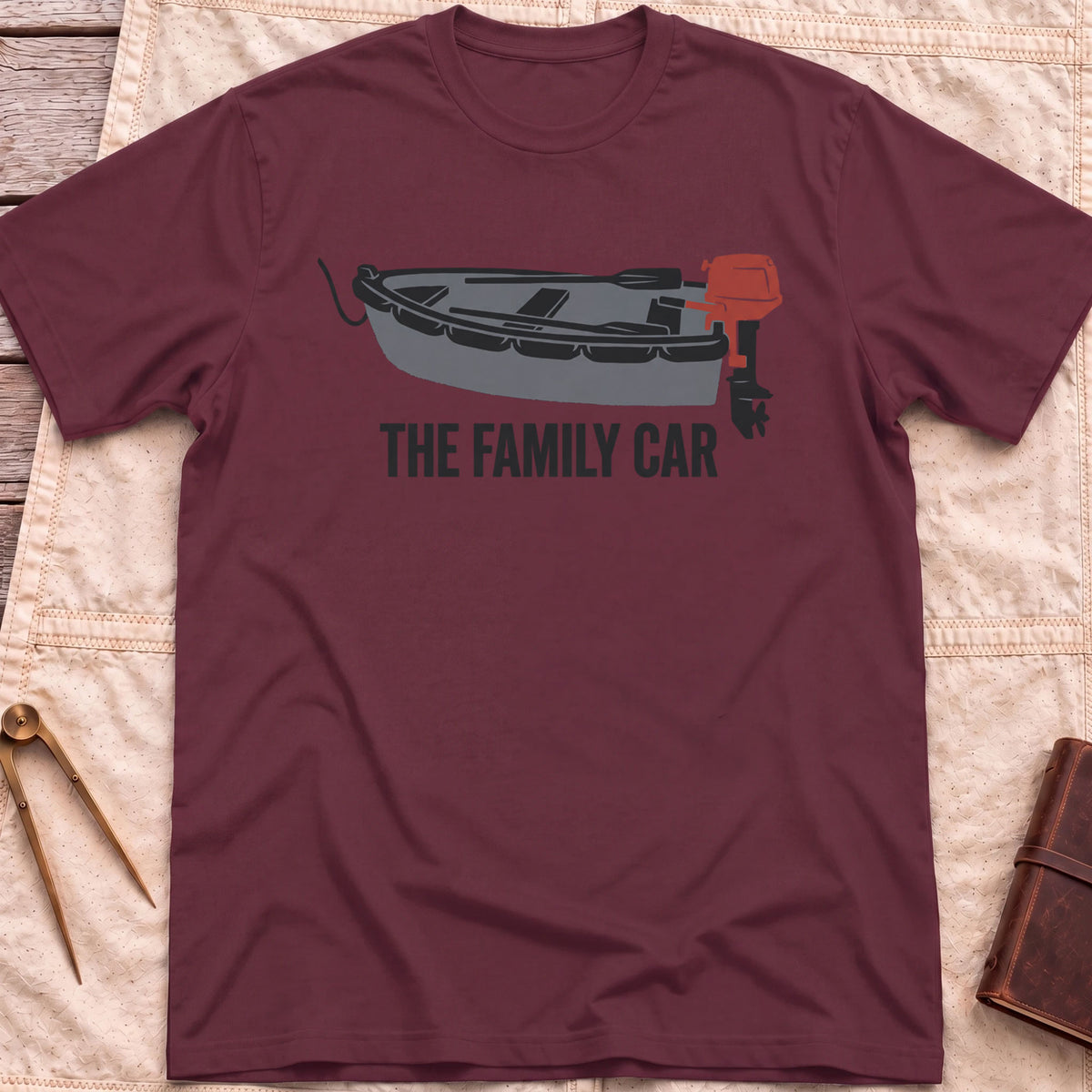 Front view of the maroon The Family Car T-Shirt with a small open boat fitted with a red outboard motor.