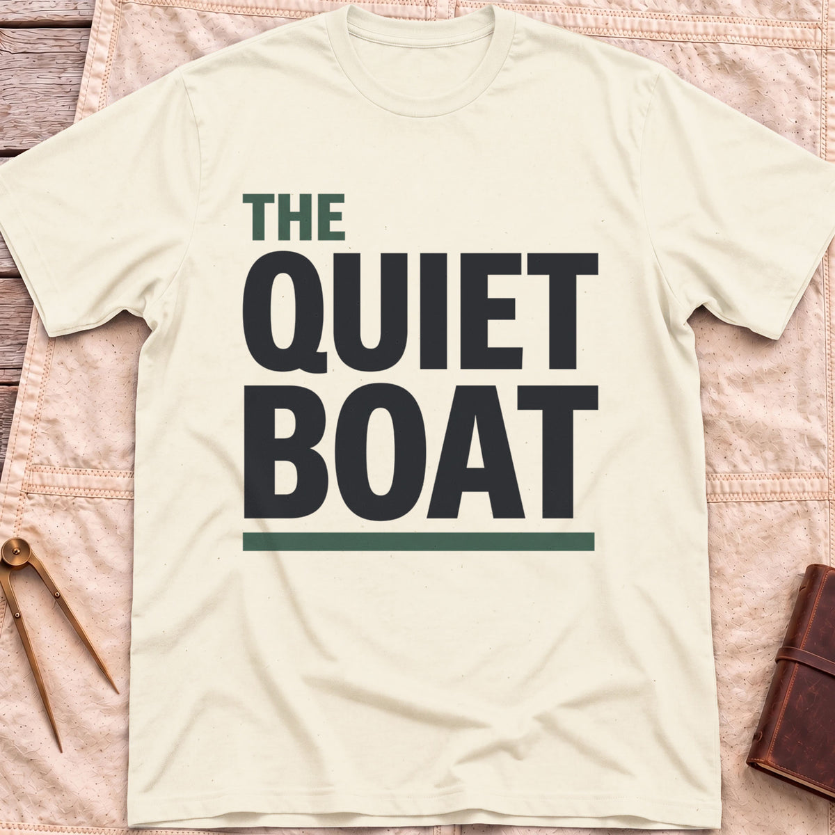 Front view of the natural The Quiet Boat T-Shirt with the words The Quiet Boat in heavy capitals above a green underline.