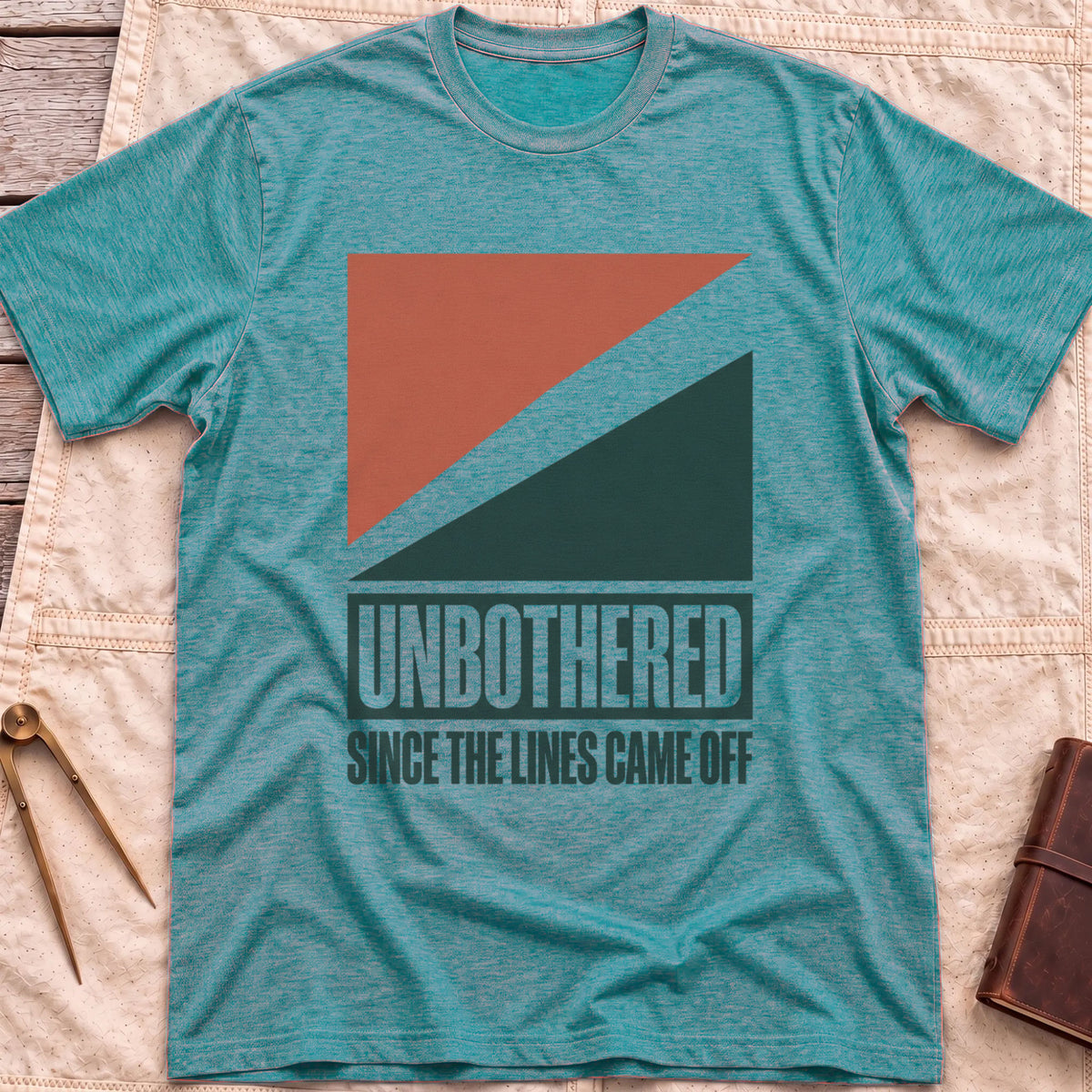 Front view of the heather galapagos blue Unbothered T-Shirt with rust-red and dark green diagonal blocks above the words Unbothered, Since the Lines Came Off.