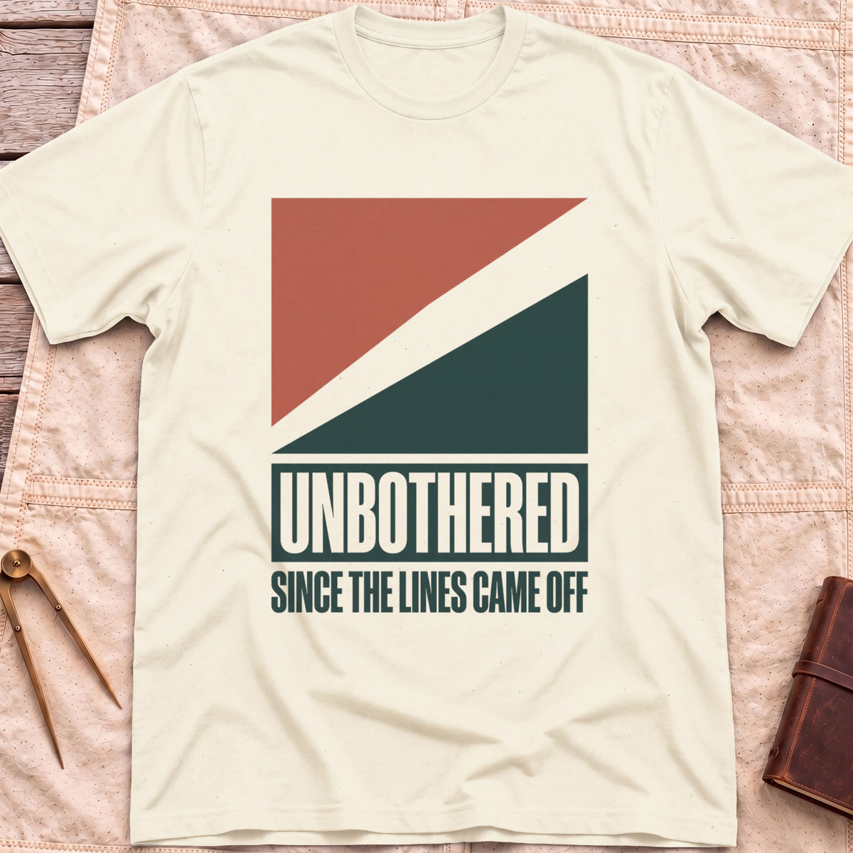 Front view of the natural Unbothered T-Shirt with rust-red and dark green diagonal blocks above the words Unbothered, Since the Lines Came Off.