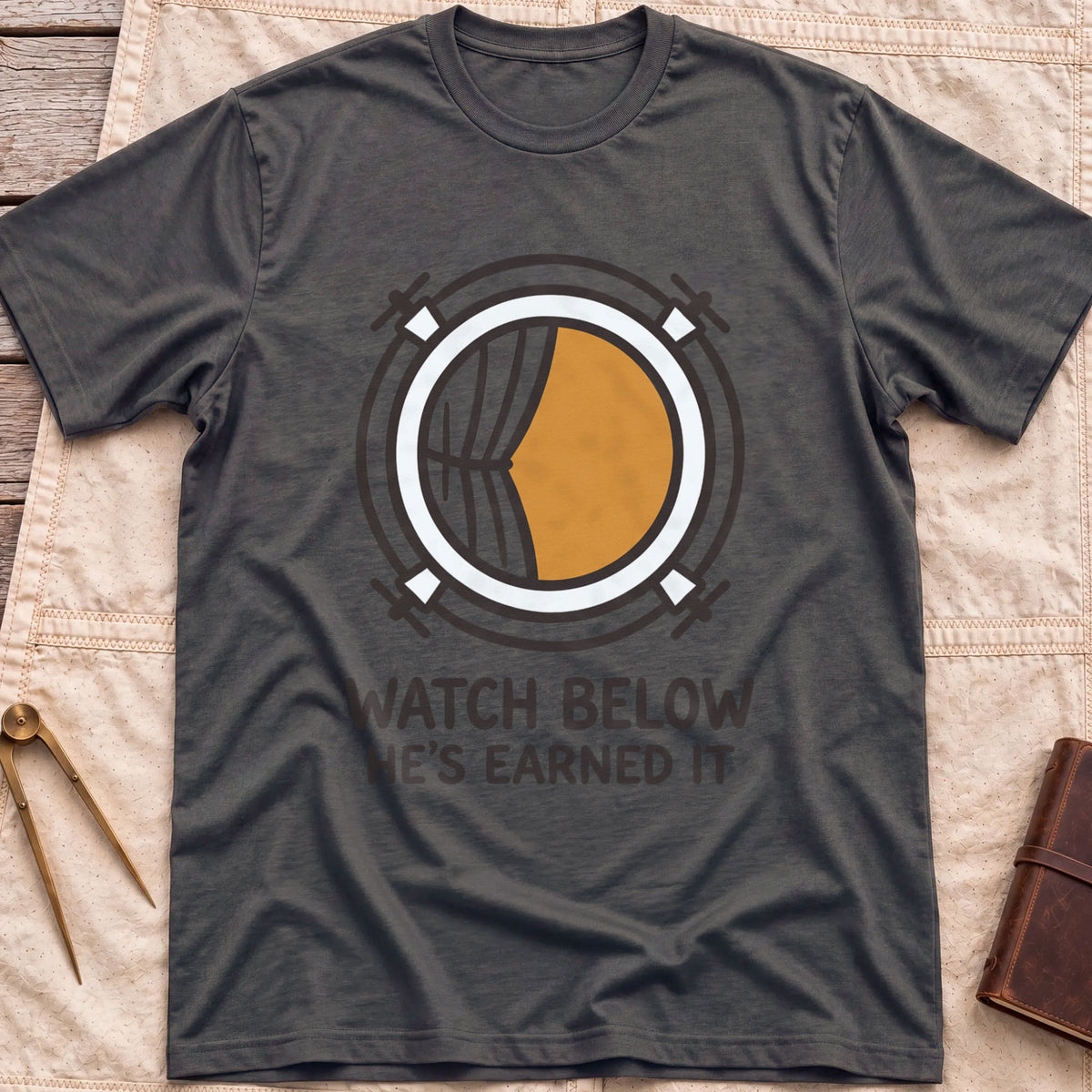 Front view of the dark heather Watch Below T-Shirt with a round porthole and a half-drawn curtain above the words He's Earned It.