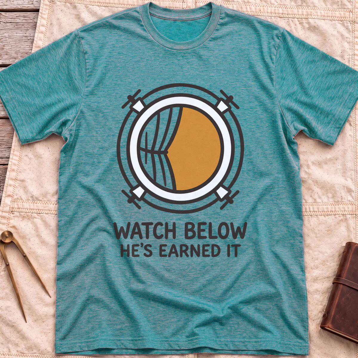 Front view of the heather galapagos blue Watch Below T-Shirt with a round porthole and a half-drawn curtain above the words He's Earned It.