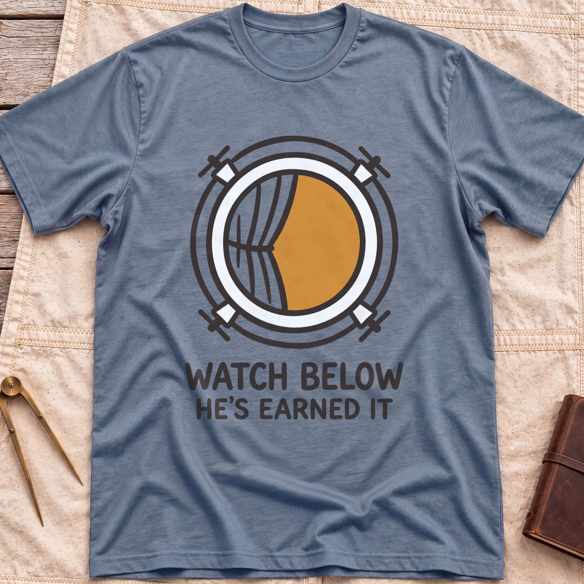 Front view of the heather indigo Watch Below T-Shirt with a round porthole and a half-drawn curtain above the words He's Earned It.