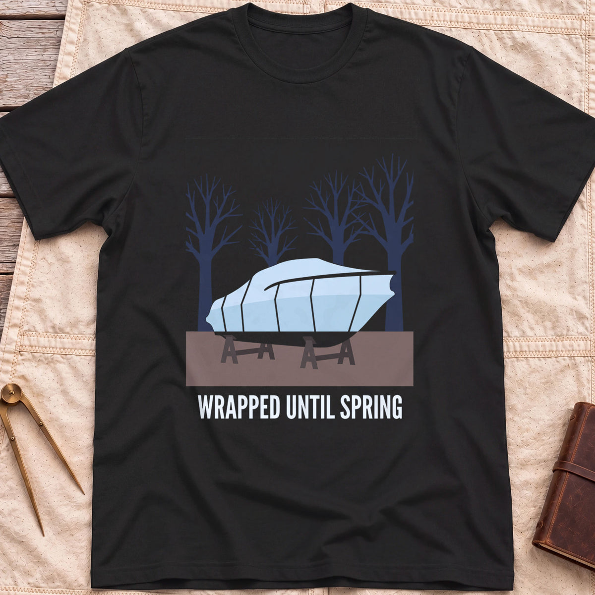 Front view of the black Wrapped Until Spring T-Shirt with a covered boat on stands in front of bare trees.