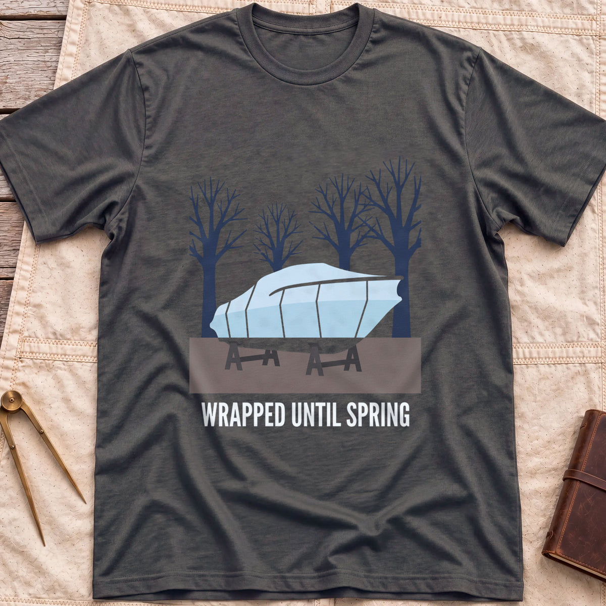 Front view of the dark heather Wrapped Until Spring T-Shirt with a covered boat on stands in front of bare trees.