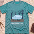 Front view of the heather galapagos blue Wrapped Until Spring T-Shirt with a covered boat on stands in front of bare trees.