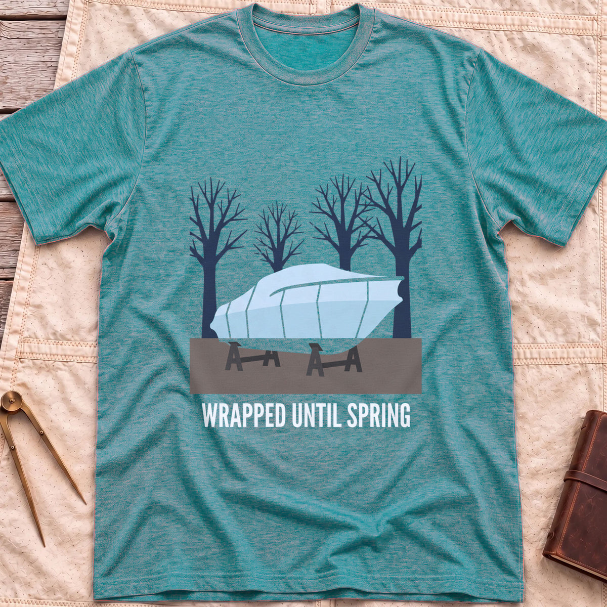Front view of the heather galapagos blue Wrapped Until Spring T-Shirt with a covered boat on stands in front of bare trees.