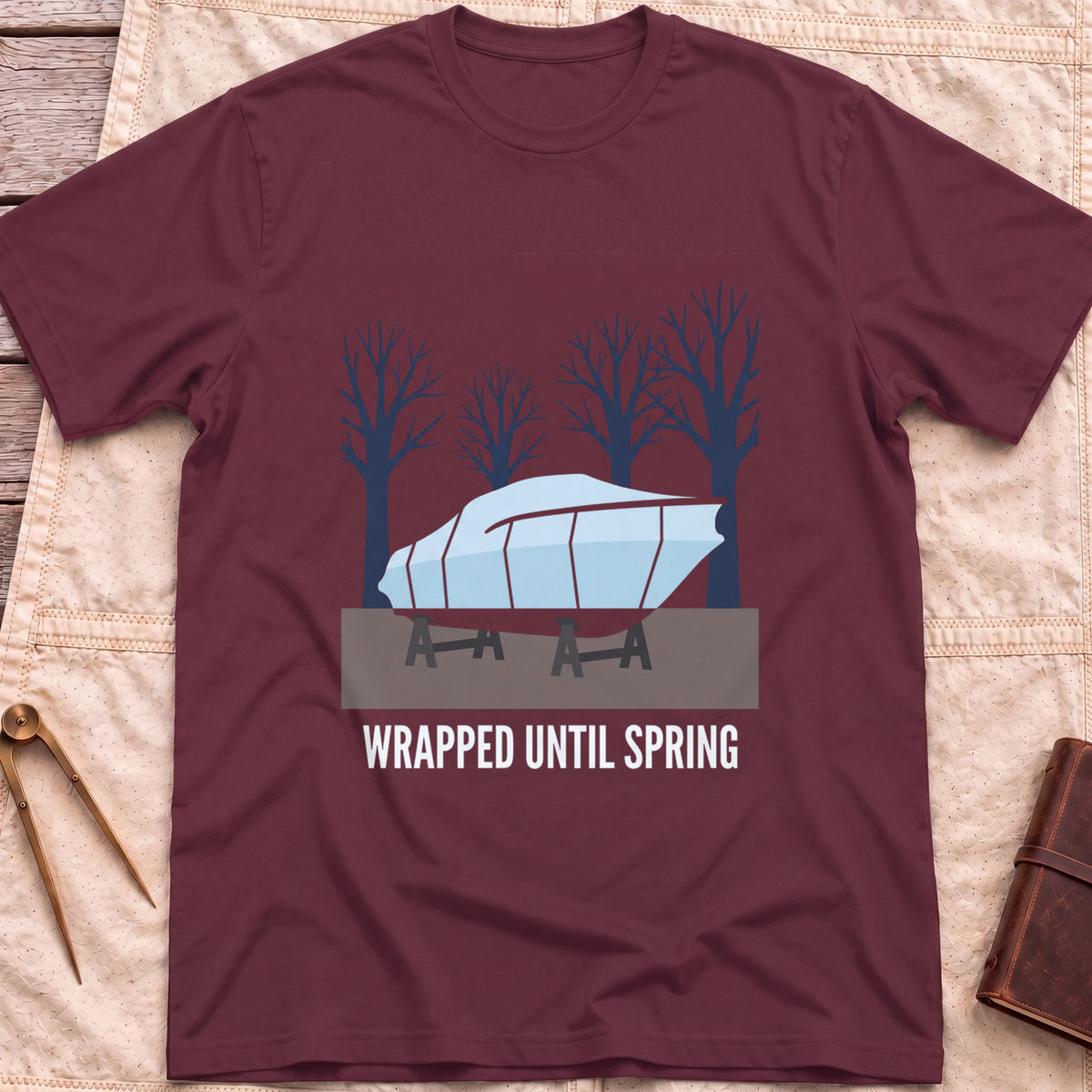 Front view of the maroon Wrapped Until Spring T-Shirt with a covered boat on stands in front of bare trees.