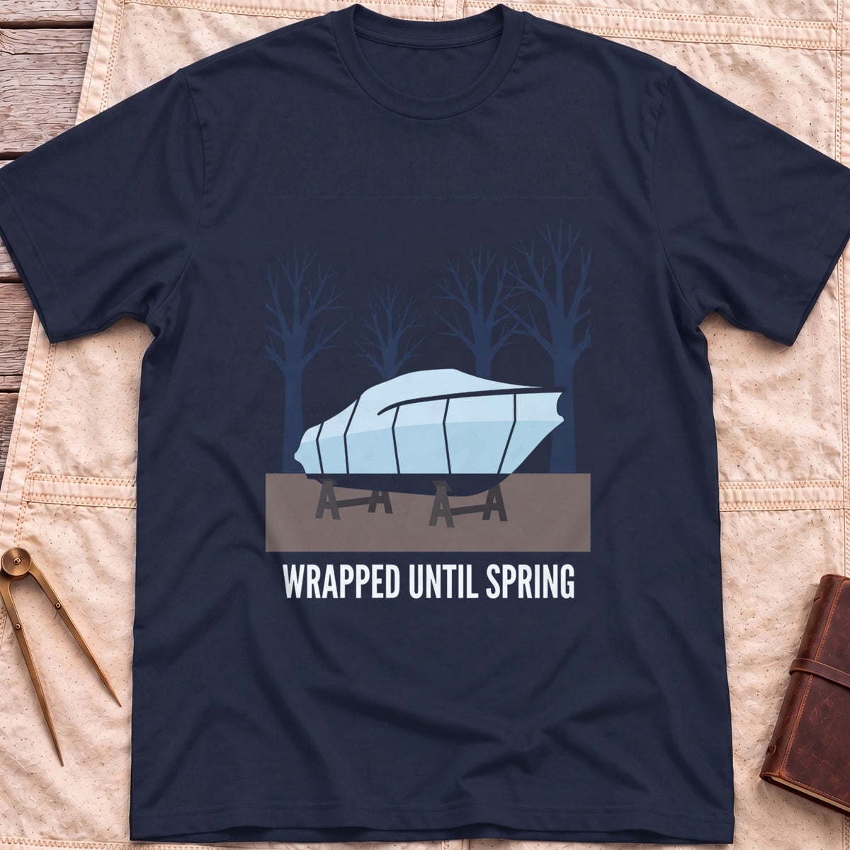 Front view of the navy Wrapped Until Spring T-Shirt with a covered boat on stands in front of bare trees.